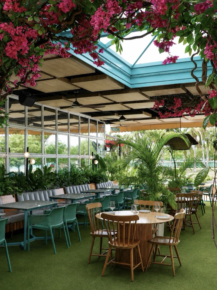 Jardín bajo techo de vidrio, con flores, palmas y mesas de madera